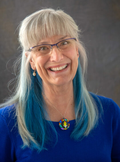 photograph of Rev. Nancy, a woman with long, straight grey hair with bright blue streaks, wearing glasses, a blue sweater, and a circular fused glass pendant with a uu chalice at the center